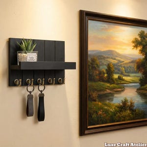 May include: A black wooden key holder with a shelf and brass hooks. A small potted plant and a glass jar with the text "hello summer" are on the shelf. Two keychains hang from the hooks. A framed landscape painting is on the right.