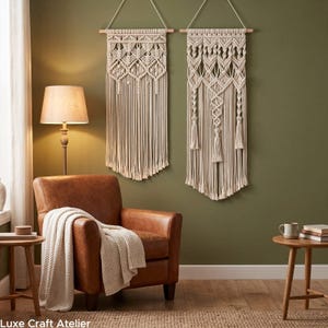 May include: Two off-white macrame wall hangings featuring intricate knotted patterns and long fringe, suspended from wooden dowels. The designs include geometric diamond shapes and vertical twisted cord elements, creating a textured appearance.