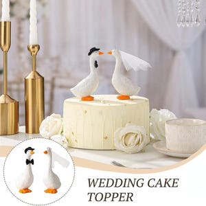 May include: Two white goose figurines serving as a wedding cake topper. One goose wears a black top hat and bow tie, while the other wears a white veil. They are positioned on a frosted cake decorated with white roses and gold leaf accents.