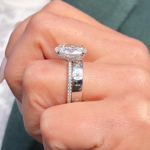 Könnte beinhalten: Nahaufnahme eines Diamant-Verlobungsrings und eines Ehering-Sets. Der Verlobungsring hat einen großen, oval geschliffenen Diamanten, der von einem Halo aus kleineren Diamanten umgeben ist. Das Ehering ist mit kleinen Diamanten und floralen Gravuren verziert.