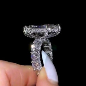 Op de afbeelding: Een sprankelende zilveren ring met een grote, rechthoekige, heldere edelsteen. De band en zetting zijn versierd met kleinere, glinsterende stenen, wat een luxueus en opvallend ontwerp creëert. De ring staat tegen een zwarte achtergrond.