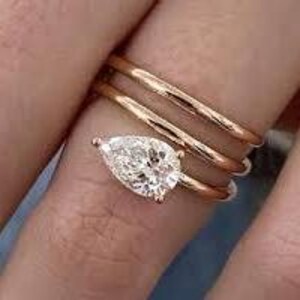 Könnte beinhalten: Nahaufnahme einer Hand mit drei goldenen Ringen. Die Ringe sind gestapelt, wobei ein birnenförmiger Diamantring im Mittelpunkt steht. Der Diamant funkelt und reflektiert das Licht. Die Ringe sind auf der Haut platziert, was den warmen Ton des Goldes hervorhebt.