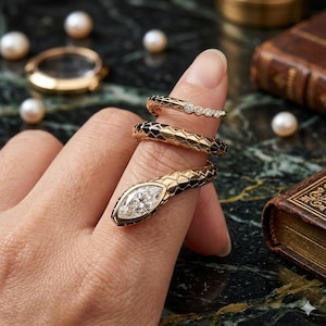 May include: Gold snake ring with a marquise-cut diamond head and black enamel detailing. The ring wraps around the finger, showcasing a unique design. Additional gold rings with diamonds are also visible, along with pearls and a vintage book.