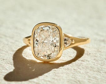 May include: A gold ring featuring a large, rectangular-cut diamond as the centerpiece. The diamond is set in a bezel setting, with smaller accent stones on either side. The band is a simple, polished gold.