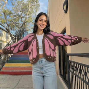 Könnte beinhalten: Ein rosa-brauner Cardigan mit Schmetterlingsflügeln und Knopfverschlüssen. Der offene Pullover hat einen V-Ausschnitt und wird mit einem weißen Oberteil und hellblauen Jeans getragen. Die Flügelspannweite des Cardigans wird gezeigt.