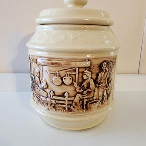 Puede incluir: Bote de galletas de cerámica color crema con tapa y escenas en relieve en relieve. El bote presenta la palabra "COOKIES" en letras en relieve. El relieve representa figuras en un entorno rústico, añadiendo encanto vintage.