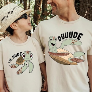 May include: Beige t-shirts with turtle graphics. One shirt says "LIL DUDE" with a cartoon turtle, and the other says "DUUUDE" with a similar turtle design. The shirts are worn by a child and an adult.