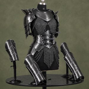 Op de afbeelding: Een set donker metalen plaatpantser met een borstplaat, schouderstukken en scheenplaten met reliëf geometrische patronen. Het pantser bevat een maliënkolder met een tailleband die wordt vastgezet met drie horizontale riemen en metalen gespen.