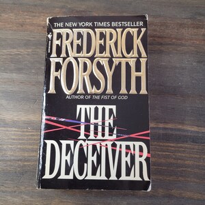 Op de afbeelding: Een zwart paperback boek met een gouden titel, "The Deceiver" van Frederick Forsyth, auteur van "The Fist of God". De omslag van het boek heeft een rood en blauw ontwerp met diagonale lijnen.