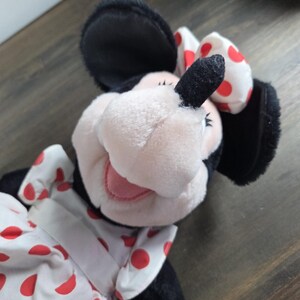 Pu&ograve; includere: Una bambola di peluche di Minnie Mouse con orecchie nere, viso rosa e un vestito bianco a pois rossi. La bambola ha un fiocco abbinato a pois rossi. La bambola &egrave; su una superficie di legno.