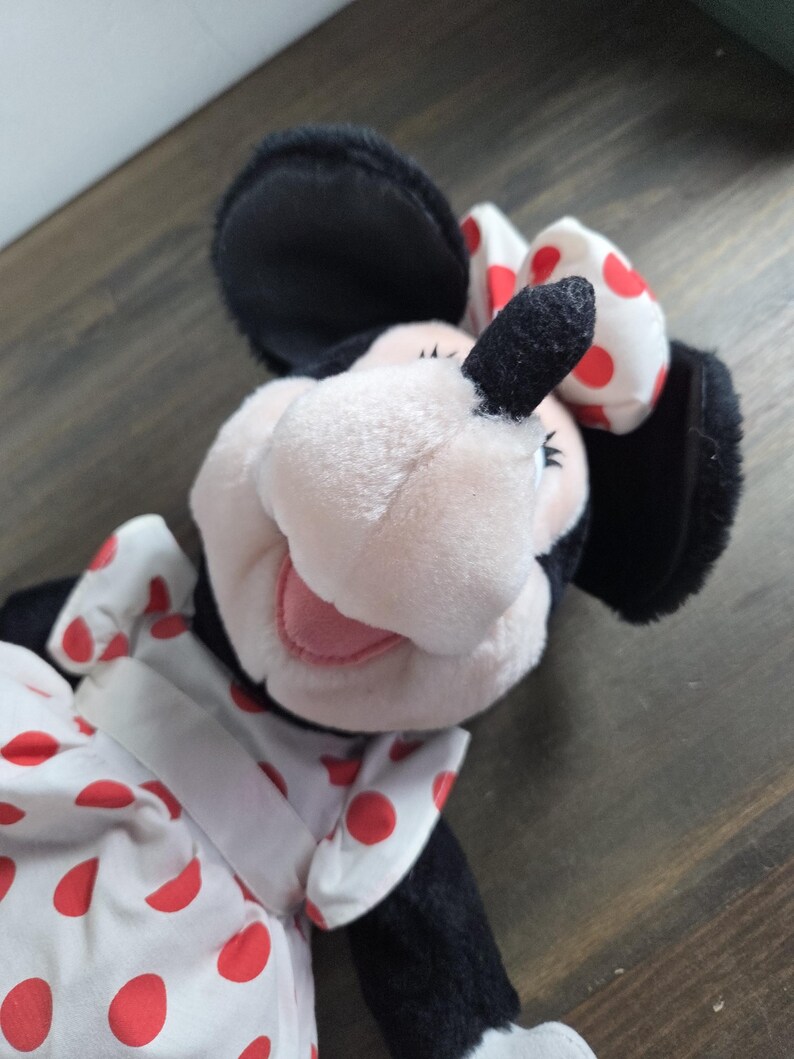 Pu&ograve; includere: Una bambola di peluche di Minnie Mouse con orecchie nere, viso rosa e un vestito bianco a pois rossi. La bambola ha un fiocco abbinato a pois rossi. La bambola &egrave; su una superficie di legno.