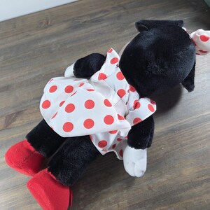 Pu&ograve; includere: Un peluche a forma di topo nero, con un vestito bianco a pois rossi e scarpe rosse. Il giocattolo ha orecchie nere e un fiocco bianco a pois rossi. &Egrave; appoggiato su una superficie di legno.