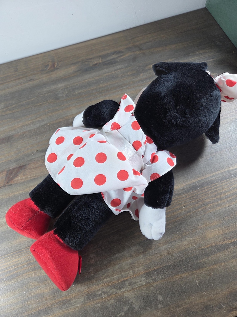 Pu&ograve; includere: Un peluche a forma di topo nero, con un vestito bianco a pois rossi e scarpe rosse. Il giocattolo ha orecchie nere e un fiocco bianco a pois rossi. &Egrave; appoggiato su una superficie di legno.