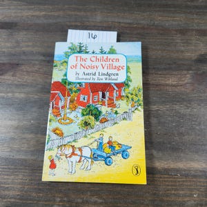 Puede incluir: Un libro infantil vintage titulado "Los Ni&ntilde;os del Pueblo Ruidoso" de Astrid Lindgren, ilustrado por Ilon Wikland. La portada presenta una ilustraci&oacute;n colorida de un pueblo con casas rojas, un carro tirado por un caballo y ni&ntilde;os jugando.