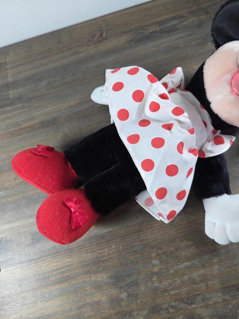 Pu&ograve; includere: Una bambola di peluche di Minnie Mouse, con un vestito bianco a pois rossi, scarpe rosse con fiocchi e gambe nere. La bambola ha un naso rosa, guanti bianchi e orecchie nere. Un classico giocattolo per bambini.