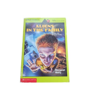 May include: A green and yellow paperback book titled "Aliens in the Family" by Margaret Mahy. The cover features an illustration of a young boy with a surprised expression, looking up at the viewer. The book is part of the Apple Paperbacks series.