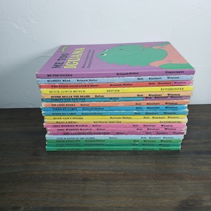 May include: A pile of children's books with vibrant covers. The top book is titled "Me Too Iguana" with a green iguana illustration. Other titles include "Scaredy Bear" and "Who Stole Alligator's Shoe!" The books are on a dark wooden surface.