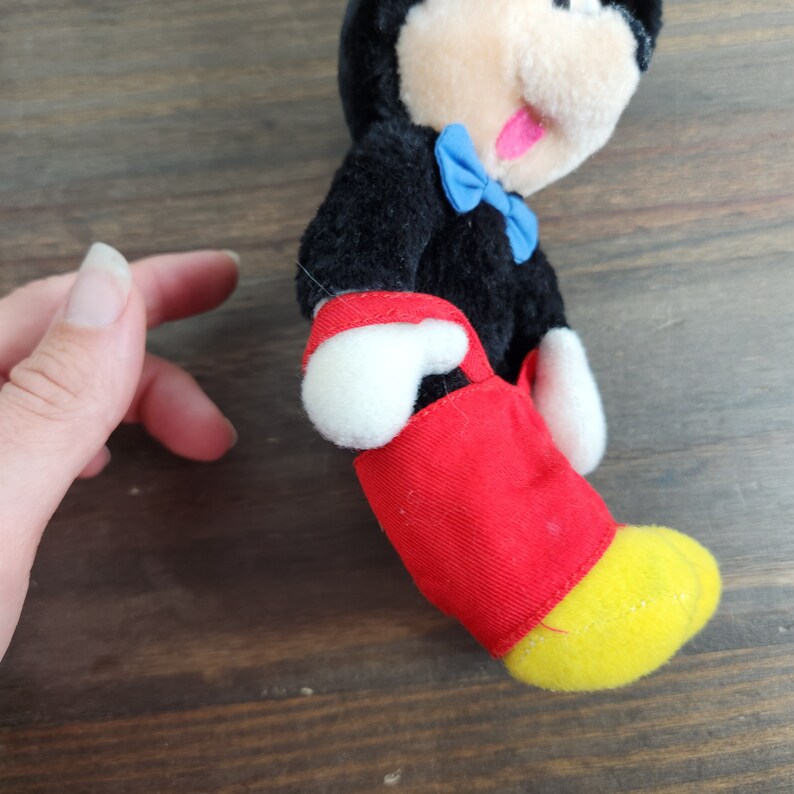 May include: A black and white plush Mickey Mouse toy wearing red pants and a blue bow tie. The toy has a yellow foot and is sitting on a wooden surface.
