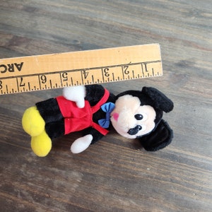 May include: A plush black and red Mickey Mouse toy with yellow shoes and a blue bow tie. The toy is lying on a wooden surface.