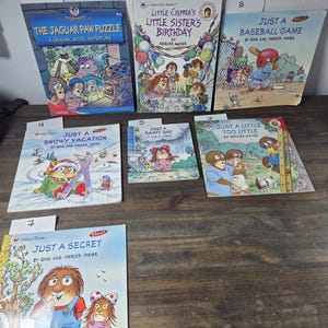 May include: A collection of children's books featuring colorful illustrations. Titles include "The Jaguar Paw Puzzle," "Little Critter's Little Sister's Birthday," and "Just a Baseball Game." The books are displayed on a wooden surface.