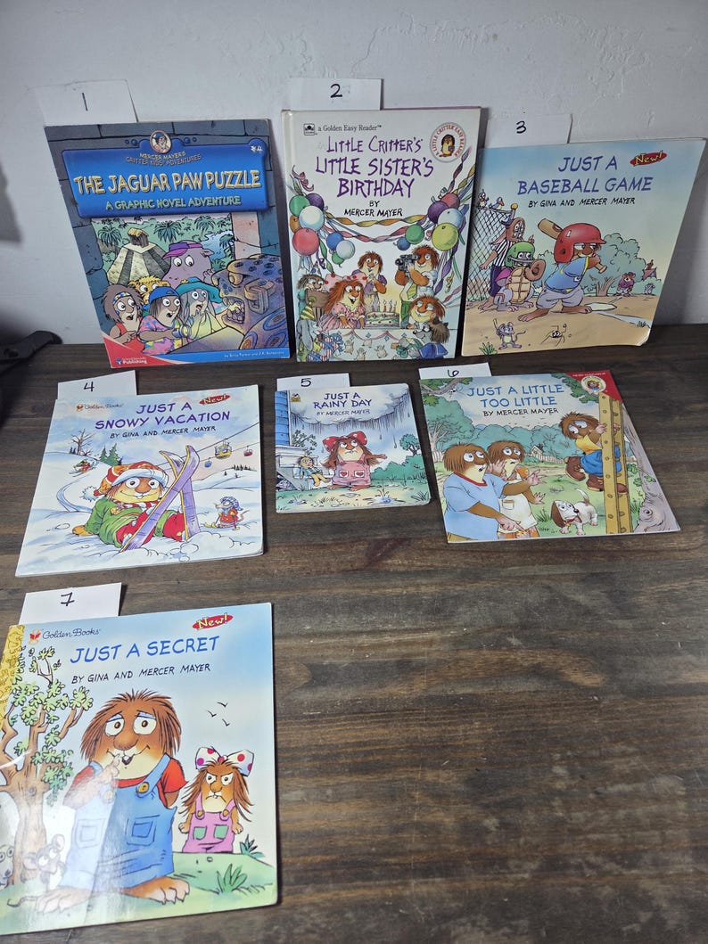 May include: A collection of children's books featuring colorful illustrations. Titles include "The Jaguar Paw Puzzle," "Little Critter's Little Sister's Birthday," and "Just a Baseball Game." The books are displayed on a wooden surface.