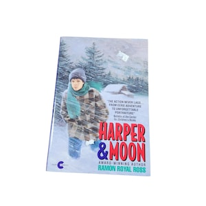 Op de afbeelding: Een boekomslag met een blauwe achtergrond en een jongen in een geruit overhemd en blauwe sjaal die door een besneeuwd bos loopt. De titel van het boek is "Harper & Moon" en de auteur is Ramon Royal Ross.