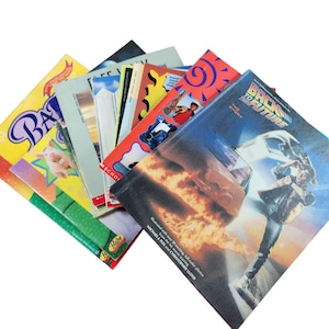 以下が含まれることがあります： 映画「バック・トゥ・ザ・フューチャー」に関する本を含む、ペーパーバックの積み重ね。表紙には「The Story」という文字が書かれており、赤、オレンジ、黄色の表紙に車と人の写真が載っている。