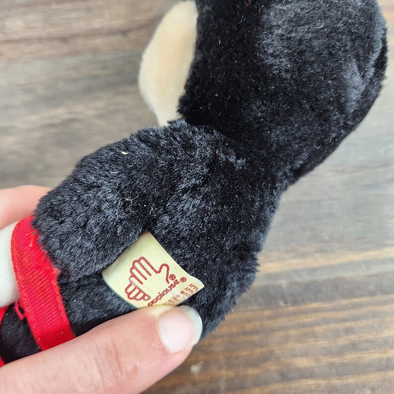 May include: Black and white plush toy with a red ribbon. The toy has a tag that says "Applique's" with a hand logo.