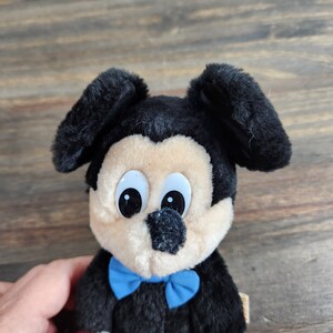 May include: A plush black and tan Mickey Mouse toy with large blue eyes and a blue bow tie. The toy is made of soft, fuzzy fabric.