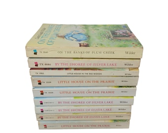 May include: A stack of eight paperback books by Laura Ingalls Wilder. The books are all different colors and have different titles, including "On the Banks of Plum Creek", "By the Shores of Silver Lake", and "Little House on the Prairie".