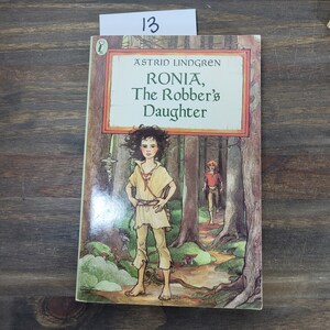Puede incluir: Una portada de libro con una ilustraci&oacute;n verde, marr&oacute;n y azul de una ni&ntilde;a de pie en un bosque. El t&iacute;tulo del libro es "Ronja, la hija del bandido" de Astrid Lindgren.