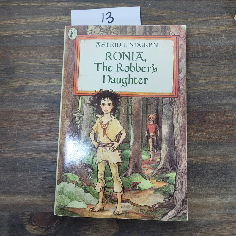 Puede incluir: Una portada de libro con una ilustraci&oacute;n verde, marr&oacute;n y azul de una ni&ntilde;a de pie en un bosque. El t&iacute;tulo del libro es "Ronja, la hija del bandido" de Astrid Lindgren.