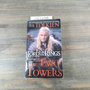 May include: A paperback book cover for "The Lord of the Rings: The Two Towers" by J.R.R. Tolkien. The cover features a photo of the character Aragorn from the movie adaptation.