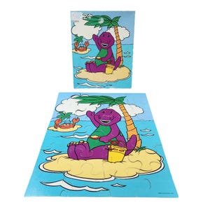 Puede incluir: Un rompecabezas de 24 piezas con Barney el dinosaurio en una isla tropical. Barney lleva una camisa verde y sostiene un cubo amarillo. Hay una palmera y un cangrejo en la isla.