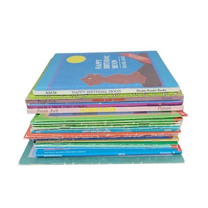 May include: A stack of children's books with colorful covers. The top book is titled "Happy Birthday, Moon" by Frank Asch. Other titles include "Milk and Cookies" and "Sand Cake". The books are in various colors, including blue, green, and pink.