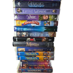 Pode incluir: Uma pilha de fitas VHS com títulos de filmes como "The Pagemaster", "Casper", "Little Women" e "E.T. The Extra-Terrestrial". As fitas têm rótulos coloridos e são empilhadas verticalmente, mostrando uma variedade de filmes para toda a família.