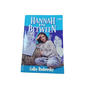 Puede incluir: Un libro de bolsillo azul y blanco titulado "Hannah in Between" con una foto de una joven en la portada. El texto "Hannah guarda un terrible secreto." está impreso debajo de la foto. El nombre del autor, Colby Rodowsky, está impreso debajo del texto.