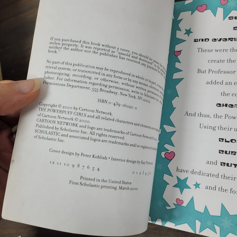 May include: A book cover with a blue background and a pink heart. The book is titled "The Powerpuff Girls" and features the characters Blossom, Bubbles, and Buttercup. The book is published by Scholastic and is part of the Cartoon Network series.