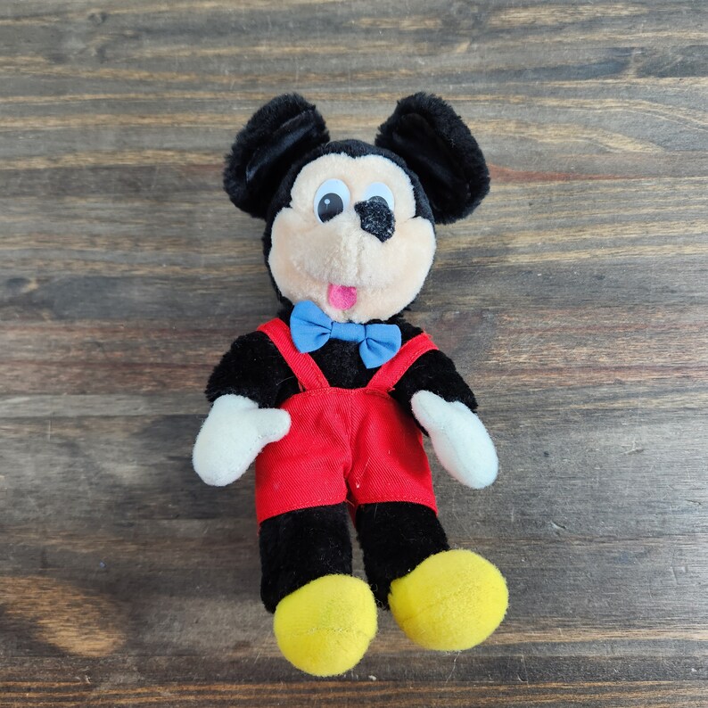 May include: A plush black Mickey Mouse toy wearing red overalls and a blue bow tie. The toy has a friendly expression with large black eyes and a pink tongue sticking out.