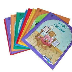 Peut inclure: Une pile de dix livres pour enfants colorés avec un livre violet sur le dessus. Le livre violet a pour titre "Wendy's Clubhouse" et présente une illustration d'un animal de dessin animé tenant un marteau. Le texte sur le livre dit "Pour avoir un ami, il faut en être un."