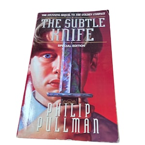 Peut inclure: Couverture du livre "The Subtle Knife" de Philip Pullman, édition spéciale. On voit un jeune homme aux yeux bleus tenant un couteau. Le titre et le nom de l'auteur sont en blanc sur fond rouge.