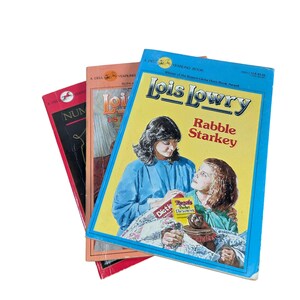 May include: A stack of three paperback books featuring the titles Rabble Starkey by Lois Lowry on the top blue and yellow cover, along with other partially visible book covers in red and orange tones, all displaying illustrated character artwork.