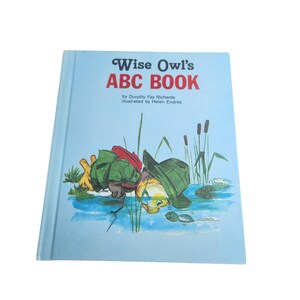 May include: A blue book titled "Wise Owl's ABC Book" by Dorothy Fay Richards, illustrated by Helen Endres. The cover shows an illustration of an owl wearing a green hat peering into a pond with a frog and a cattail.