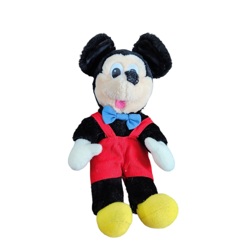 May include: A black and white plush Mickey Mouse toy wearing red overalls and a blue bow tie. The toy has a big smile and large, round eyes.
