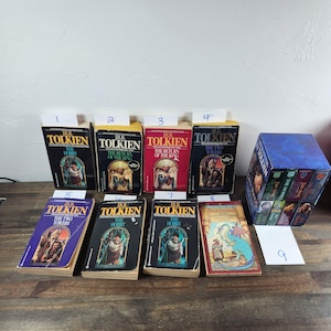 May include: A collection of nine paperback books by J.R.R. Tolkien, including "The Hobbit", "The Fellowship of the Ring", "The Two Towers", and "The Return of the King". The books are arranged in a row on a wooden surface.