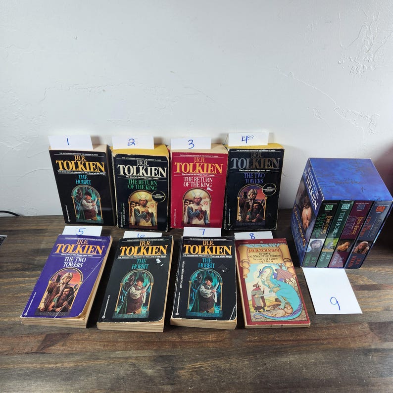May include: A collection of nine paperback books by J.R.R. Tolkien, including "The Hobbit", "The Fellowship of the Ring", "The Two Towers", and "The Return of the King". The books are arranged in a row on a wooden surface.