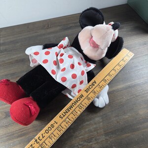 Pu&ograve; includere: Una bambola di peluche di Minnie Mouse, con pelliccia nera, un vestito bianco a pois rossi e scarpe rosse con fiocchi. La bambola ha naso e orecchie rosa. Un righello di legno &egrave; accanto alla bambola.