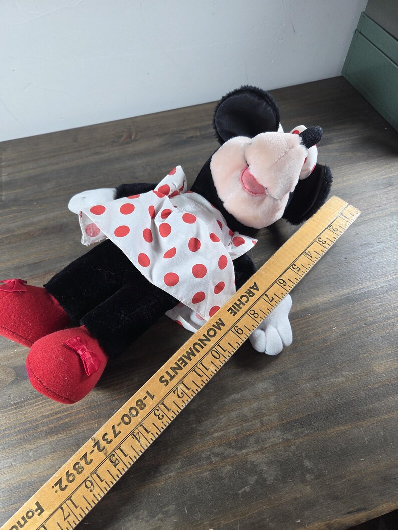 Pu&ograve; includere: Una bambola di peluche di Minnie Mouse, con pelliccia nera, un vestito bianco a pois rossi e scarpe rosse con fiocchi. La bambola ha naso e orecchie rosa. Un righello di legno &egrave; accanto alla bambola.