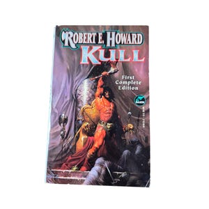 Könnte beinhalten: Ein Vintage-Taschenbuch mit dem Titel "Robert E. Howard Kull" mit einer Fantasy-Illustration. Das Cover zeigt eine muskulöse Figur, die eine Waffe hält und auf einem Thron sitzt, mit dem Text "First Complete Edition".