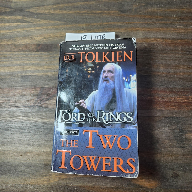 May include: A paperback book cover for "The Lord of the Rings: The Two Towers" by J.R.R. Tolkien. The cover features a photo of Gandalf the Grey, a wizard from the book series, with a white beard and a staff. The book cover is orange, red, and white.